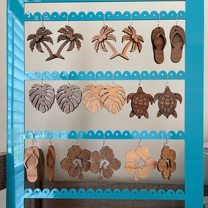 May include: A turquoise wooden earring display stand with multiple rows of wooden earrings. The earrings are shaped like palm trees, monstera leaves, flip flops, turtles, and hibiscus flowers.