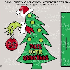 Może przedstawiać: Zielone choinka z ręką Grincha trzymającą czerwoną ozdobę z numerem 25. Na choince widnieje napis "Days Until Grinchmas". Choinka ma 19 cali wysokości, 15 cali szerokości i 3 cale głębokości.