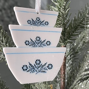 Puede incluir: Adornos navideños de cerámica blanca con forma de cuencos, con diseños florales azules y ribetes azules. Cada cuenco, de diferente tamaño, está suspendido por una cinta blanca. Cuelgan de un árbol de Navidad escarchado.