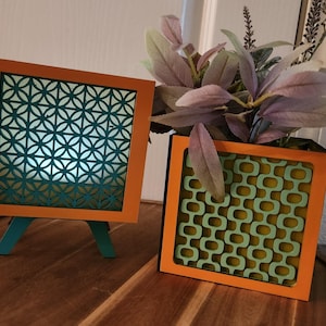 May include: Two decorative square wooden boxes with orange frames. One box has a teal geometric pattern, and the other has a teal retro pattern. The boxes are on a wooden surface with a plant in the background.