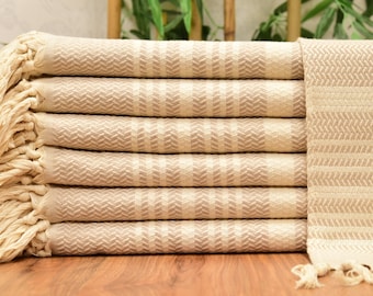 May include: A stack of beige-colored Turkish towels with a geometric pattern. The towels have a fringed edge and are folded neatly. The pattern is a series of vertical lines with a chevron design. The towels are stacked on a wooden surface.