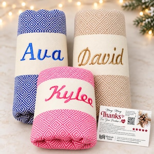 May include: Three rolled towels featuring a diamond pattern in blue, beige, and pink. Each towel has a cream-colored band embroidered with the names Ava in blue, David in gold, and Kylee in pink. A small white thank you card is included.