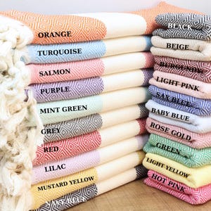 May include: A stack of Turkish towels in various colors, including orange, turquoise, salmon, purple, mint green, grey, red, lilac, and mustard yellow. Each towel features a diamond pattern and is labeled with its color name. Additional colors include black, beige, burgundy, light pink, sax blue, baby blue, rose gold, green, light yellow, and hot pink.