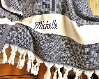 Può includere: Una coperta a fantasia blu navy e bianca con il nome "Michelle" ricamato in corsivo. La coperta presenta un motivo a rombi e nappe bianche lungo il bordo. La coperta è drappeggiata su una superficie di legno.