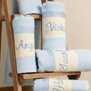 May include: Light blue and cream-coloured personalised towels with embroidered names. The towels are stacked on a wooden shelf. The towels have a diamond pattern and striped accents.