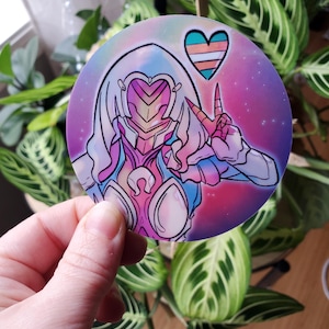 May include: A round sticker featuring a stylized illustration of a figure with a helmet and a heart with the transgender pride flag colors. The background is a gradient of pink, purple, and blue with stars. The figure is making a peace sign.