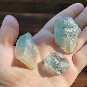 May include: Three raw aquamarine crystals of different sizes, held in an open hand. The crystals are pale blue and translucent, with natural, rough textures. The background is a wooden surface.