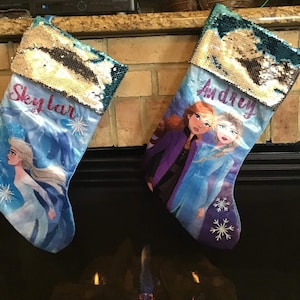 May include: Two blue and purple Christmas stockings with a Frozen theme. One stocking has the name "Skylar" embroidered on it and the other has the name "Audrey" embroidered on it.