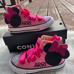 May include: Pink high-top Converse sneakers with a black Minnie Mouse design and pink bow. The shoes have a white sole and are decorated with rhinestones. The name "ARRI" is written on the side of the shoes.