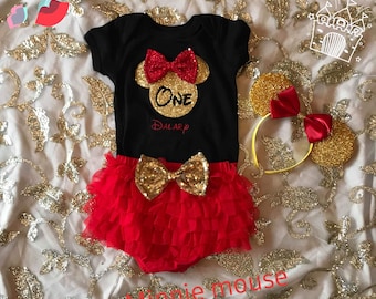 May include: A black bodysuit with a red bow and gold glitter Minnie Mouse head with the word "ONE" in gold glitter. The bodysuit is paired with a red tulle skirt with a gold glitter bow. A yellow headband with gold glitter Minnie Mouse ears is also included.