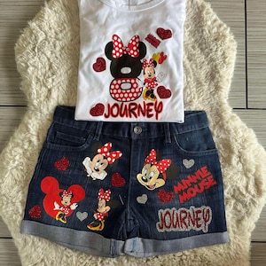 Peut inclure: Ensemble compos&eacute; d'un d&eacute;bardeur blanc et d'un short en jean avec des motifs de Minnie Mouse. Le haut pr&eacute;sente un grand motif de Minnie Mouse avec le mot "JOURNEY". Le short comporte plusieurs motifs de Minnie Mouse et les mots "MINNIE MOUSE JOURNEY".
