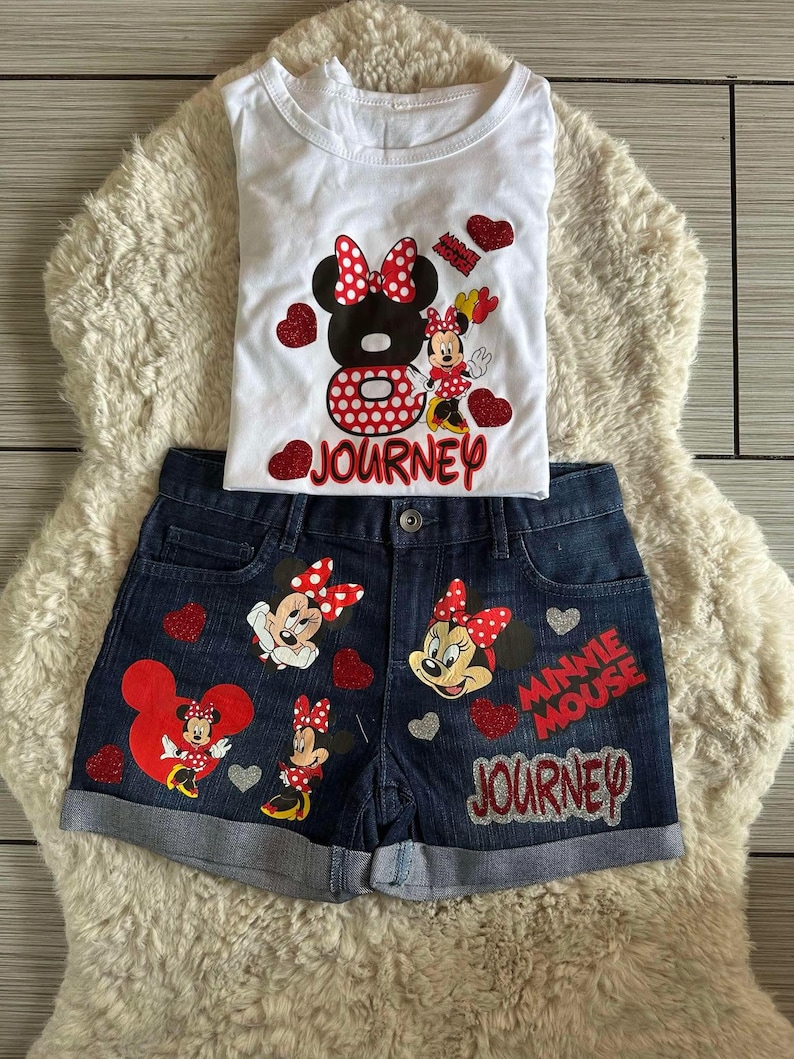 Peut inclure: Ensemble compos&eacute; d'un d&eacute;bardeur blanc et d'un short en jean avec des motifs de Minnie Mouse. Le haut pr&eacute;sente un grand motif de Minnie Mouse avec le mot "JOURNEY". Le short comporte plusieurs motifs de Minnie Mouse et les mots "MINNIE MOUSE JOURNEY".