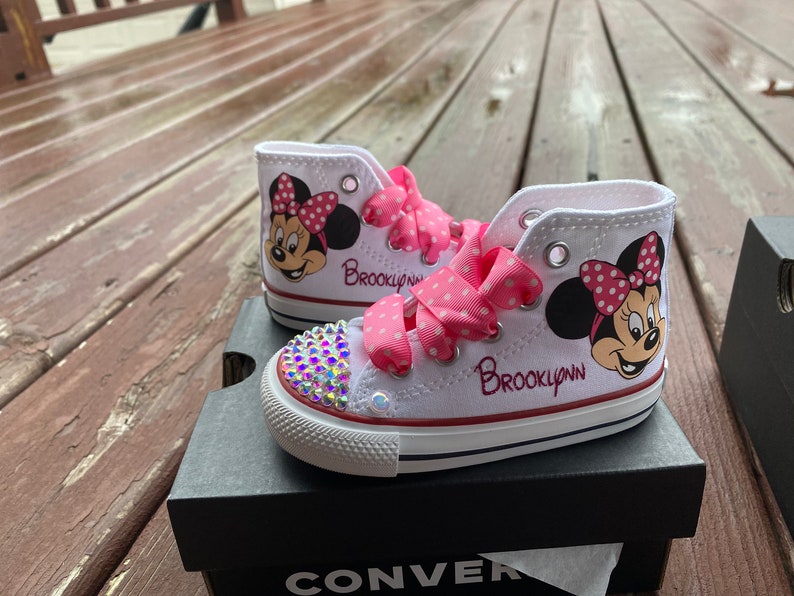 May include: White high-top sneakers with pink polka dot ribbon laces and a Minnie Mouse design. The sneakers are decorated with rhinestones and the name "Brooklynn" is written on the side.