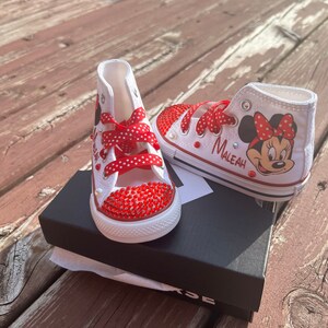 Peut inclure: Baskets montantes blanches avec des lacets rouges &agrave; pois et des ornements en strass rouges. Une chaussure pr&eacute;sente un dessin de Minnie Mouse et le nom "Maleah". Les chaussures sont sur une bo&icirc;te noire, pour enfant.