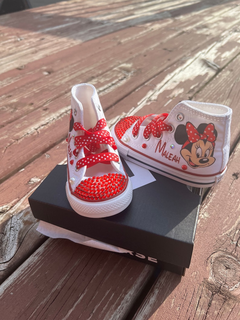 Peut inclure: Baskets montantes blanches avec des lacets rouges &agrave; pois et des ornements en strass rouges. Une chaussure pr&eacute;sente un dessin de Minnie Mouse et le nom "Maleah". Les chaussures sont sur une bo&icirc;te noire, pour enfant.