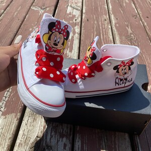 Puede incluir: Zapatillas blancas de ca&ntilde;a alta con cordones de lunares rojos y blancos y un dise&ntilde;o de Minnie Mouse en el lateral. El nombre "Alaina" est&aacute; escrito en una zapatilla.