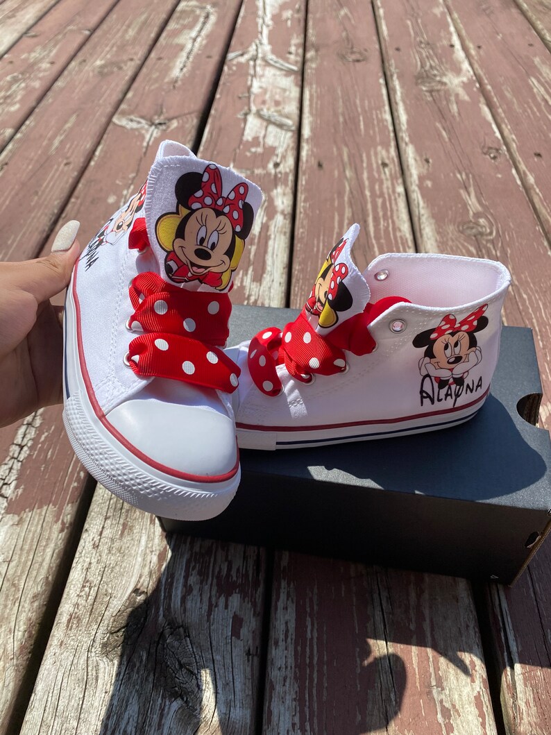 Puede incluir: Zapatillas blancas de ca&ntilde;a alta con cordones de lunares rojos y blancos y un dise&ntilde;o de Minnie Mouse en el lateral. El nombre "Alaina" est&aacute; escrito en una zapatilla.