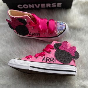May include: Pink high-top Converse sneakers with a black and pink Minnie Mouse design and rhinestones. The shoes are personalized with the name "ARRI" on each shoe.