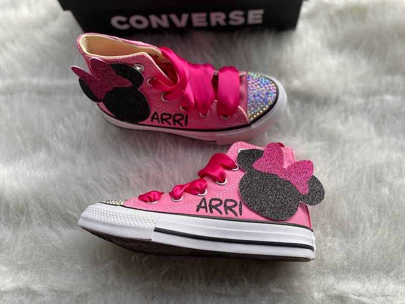 May include: Pink high-top Converse sneakers with a black and pink Minnie Mouse design and rhinestones. The shoes are personalized with the name "ARRI" on each shoe.