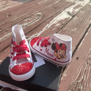 Peut inclure: Baskets montantes blanches avec des strass rouges et des lacets rouges &agrave; pois. Une chaussure pr&eacute;sente un dessin de Minnie Mouse et le nom "Maleah". Les chaussures sont sur une bo&icirc;te noire, sur un fond en bois.