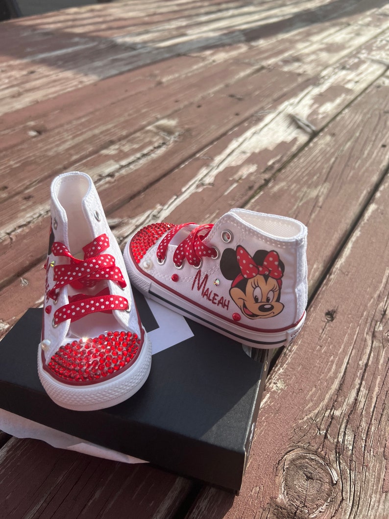 Peut inclure: Baskets montantes blanches avec des strass rouges et des lacets rouges &agrave; pois. Une chaussure pr&eacute;sente un dessin de Minnie Mouse et le nom "Maleah". Les chaussures sont sur une bo&icirc;te noire, sur un fond en bois.