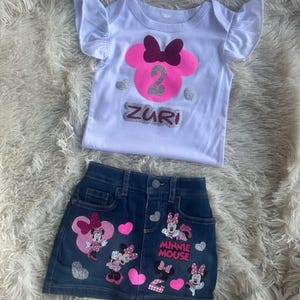 Puede incluir: Una camiseta blanca con una cabeza de Minnie Mouse rosa con un lazo burdeos y el n&uacute;mero 2 en purpurina plateada. La camiseta dice "Zuri" en purpurina plateada. Una falda vaquera con corazones rosas y aplicaciones de Minnie Mouse. La falda dice "Minnie Mouse" en purpurina plateada.