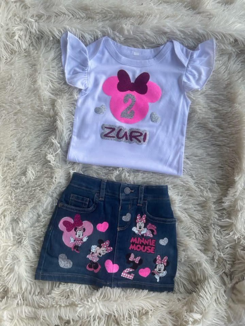 Puede incluir: Una camiseta blanca con una cabeza de Minnie Mouse rosa con un lazo burdeos y el n&uacute;mero 2 en purpurina plateada. La camiseta dice "Zuri" en purpurina plateada. Una falda vaquera con corazones rosas y aplicaciones de Minnie Mouse. La falda dice "Minnie Mouse" en purpurina plateada.