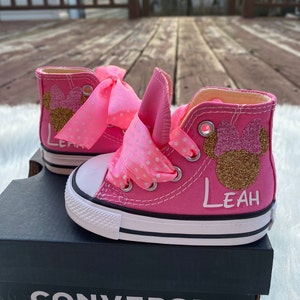 Puede incluir: Zapatillas Converse de ca&ntilde;a alta de color rosa con un lazo de cinta de lunares rosa y blanco, un dise&ntilde;o de Minnie Mouse con purpurina dorada y el nombre "Leah" escrito en el lateral.
