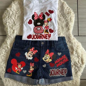 Peut inclure: Ensemble compos&eacute; d'un d&eacute;bardeur blanc et d'un short en jean avec des motifs de Minnie Mouse. Le haut pr&eacute;sente un grand motif de Minnie Mouse avec le mot "JOURNEY". Le short a plusieurs motifs de Minnie Mouse et les mots "MINNIE MOUSE JOURNEY".