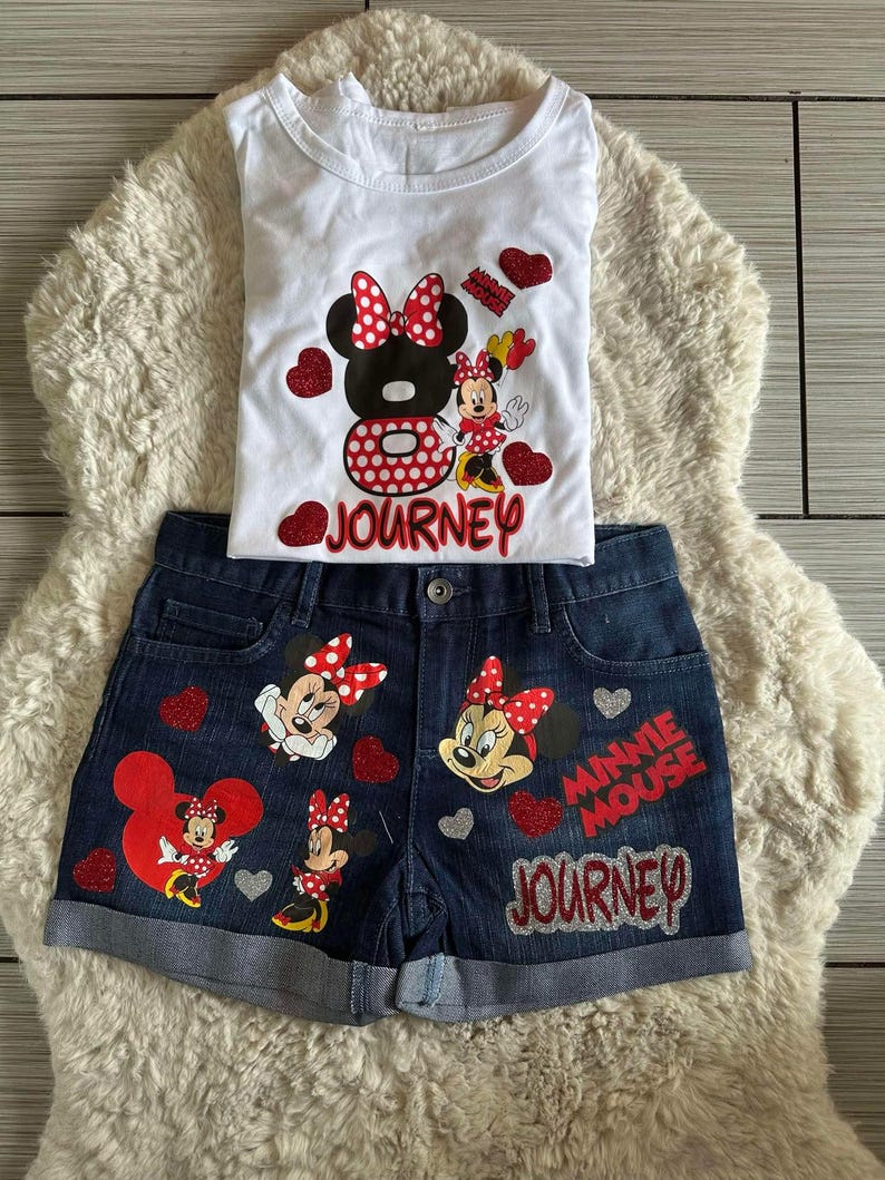 Peut inclure: Ensemble compos&eacute; d'un d&eacute;bardeur blanc et d'un short en jean avec des motifs de Minnie Mouse. Le haut pr&eacute;sente un grand motif de Minnie Mouse avec le mot "JOURNEY". Le short a plusieurs motifs de Minnie Mouse et les mots "MINNIE MOUSE JOURNEY".