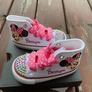 May include: White high-top sneakers with pink polka dot ribbon laces and a Minnie Mouse design. The shoes are decorated with rhinestones and the name "Brooklynn" is written on the side.