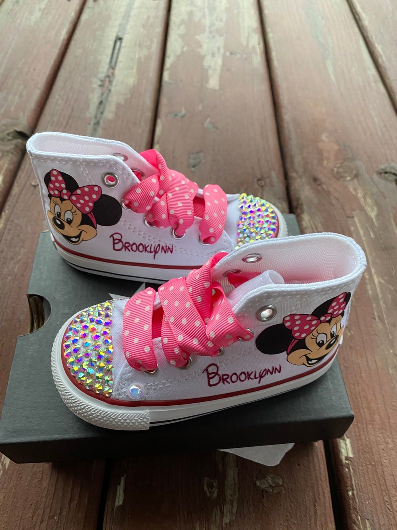 May include: White high-top sneakers with pink polka dot ribbon laces and a Minnie Mouse design. The shoes are decorated with rhinestones and the name "Brooklynn" is written on the side.