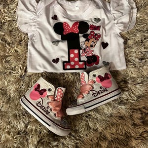 K&ouml;nnte beinhalten: Ein wei&szlig;er Baby-Body und passende High-Top-Sneaker mit Minnie-Maus-Motiv. Der Body hat eine gro&szlig;e "1" mit Polka-Dots und Minnie-Maus-Grafiken. Die Sneaker sind wei&szlig; mit rosa Schleifen und dem Namen "Dezire".