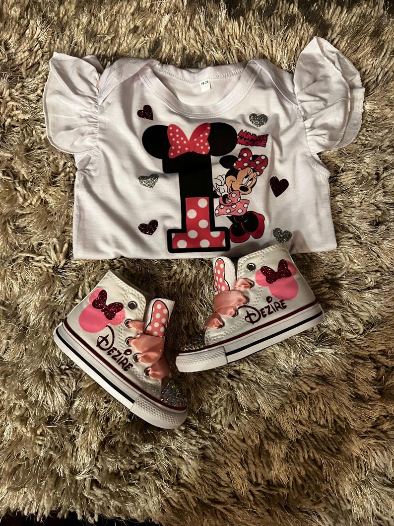 K&ouml;nnte beinhalten: Ein wei&szlig;er Baby-Body und passende High-Top-Sneaker mit Minnie-Maus-Motiv. Der Body hat eine gro&szlig;e "1" mit Polka-Dots und Minnie-Maus-Grafiken. Die Sneaker sind wei&szlig; mit rosa Schleifen und dem Namen "Dezire".