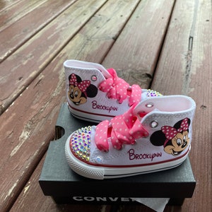May include: White high-top sneakers with pink polka dot ribbon laces and a Minnie Mouse design. The sneakers are personalized with the name "Brooklyn" in red lettering. The sneakers are decorated with rhinestones.
