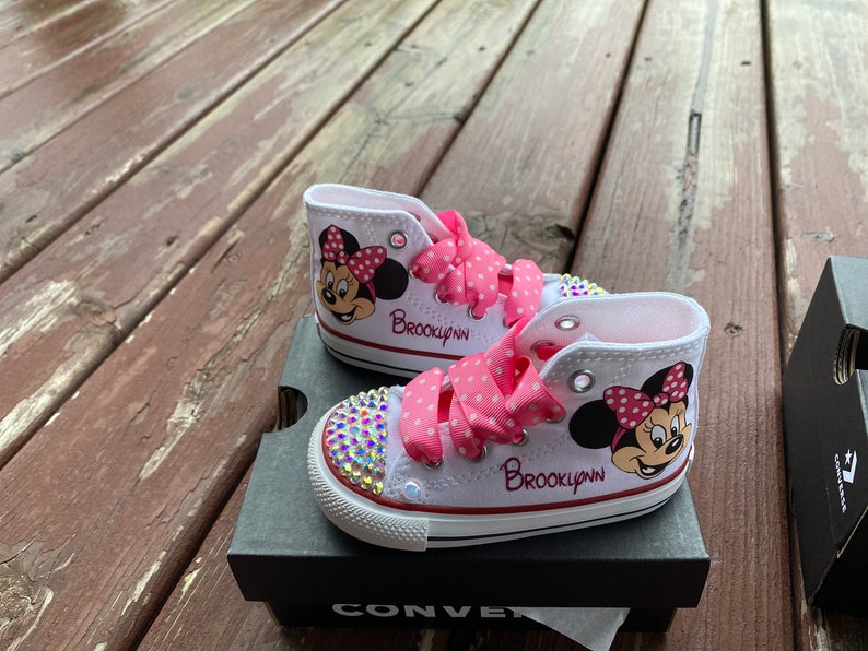 May include: White high-top sneakers with pink polka dot ribbon laces and a Minnie Mouse design. The sneakers are personalized with the name "Brooklyn" in red lettering. The sneakers are decorated with rhinestones.