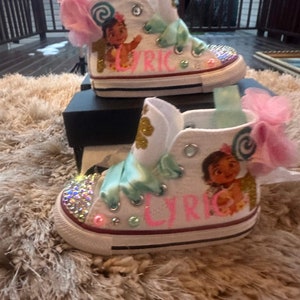 May include: White high-top trainers with a pink and green bow, glitter, and a Moana character design. The shoes are personalised with the name "Lyric".