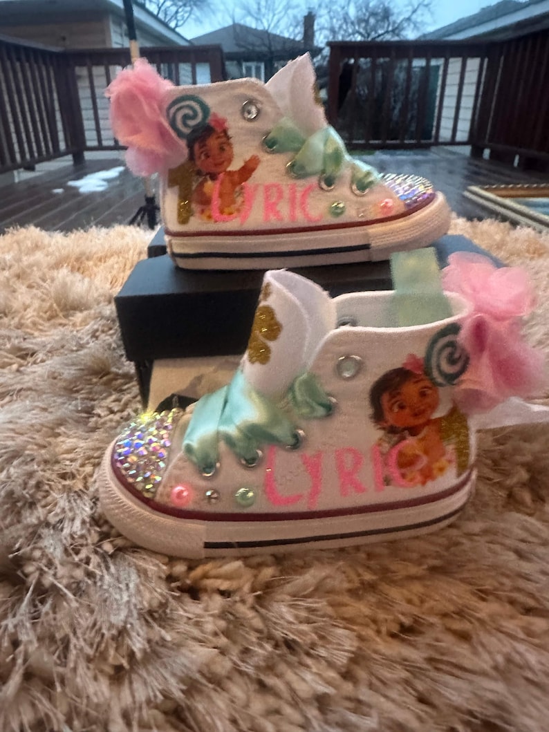May include: White high-top trainers with a pink and green bow, glitter, and a Moana character design. The shoes are personalised with the name "Lyric".