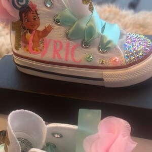 May include: White high-top trainers with a pink flower, green ribbon, and a cartoon image of Moana from Disney's Moana. The trainers are decorated with rhinestones and the name "Lyric" is written in pink glitter.