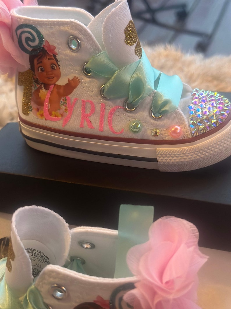 May include: White high-top trainers with a pink flower, green ribbon, and a cartoon image of Moana from Disney's Moana. The trainers are decorated with rhinestones and the name "Lyric" is written in pink glitter.