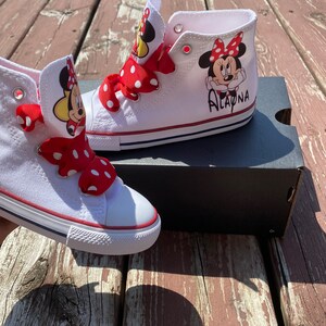 Puede incluir: Zapatillas blancas de ca&ntilde;a alta con cordones de cinta de lunares rojos y blancos. Las zapatillas tienen una imagen de dibujos animados de Minnie Mouse con el nombre "Alayna" escrito en el lateral.