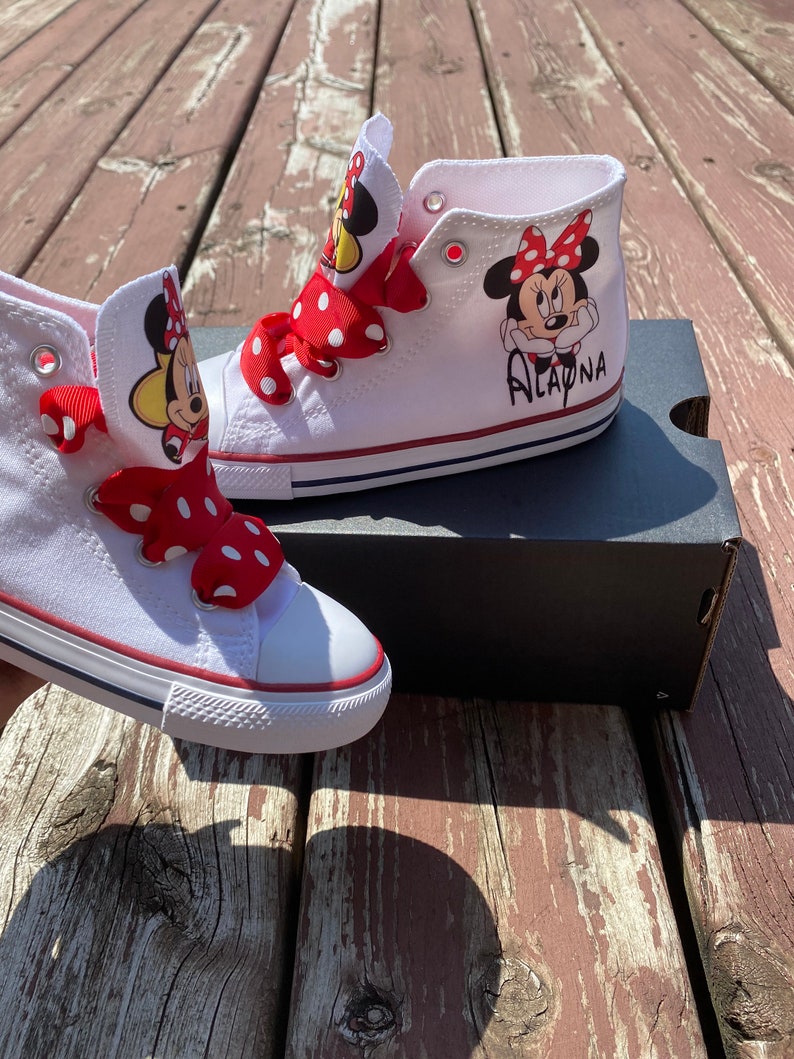 Puede incluir: Zapatillas blancas de ca&ntilde;a alta con cordones de cinta de lunares rojos y blancos. Las zapatillas tienen una imagen de dibujos animados de Minnie Mouse con el nombre "Alayna" escrito en el lateral.