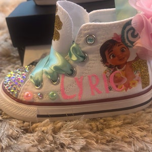 May include: White canvas high-top trainers with a pink glitter design that says "Lyric" and a Moana character graphic. The shoes have a green ribbon and a pink flower.