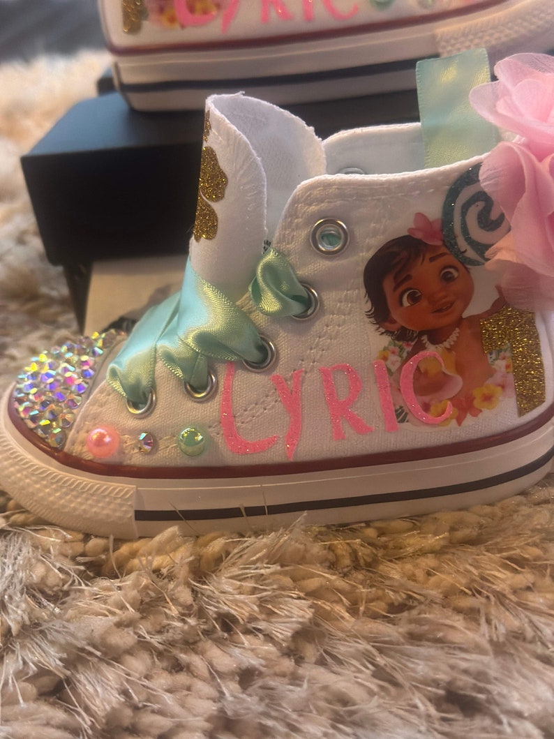 May include: White canvas high-top trainers with a pink glitter design that says "Lyric" and a Moana character graphic. The shoes have a green ribbon and a pink flower.