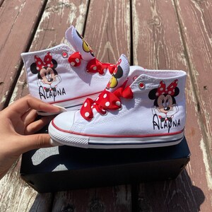 Puede incluir: Zapatillas blancas de ca&ntilde;a alta con lazos rojos de lunares blancos y un dise&ntilde;o de Minnie Mouse. Las zapatillas est&aacute;n personalizadas con el nombre "Alayna".