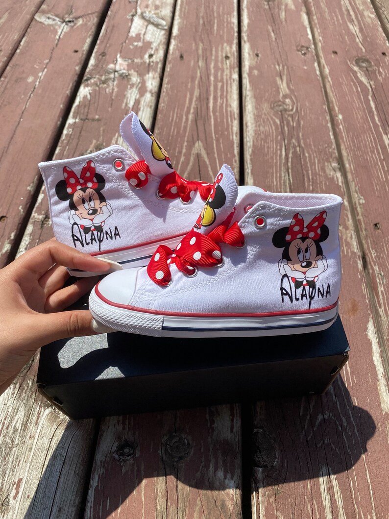 Puede incluir: Zapatillas blancas de ca&ntilde;a alta con lazos rojos de lunares blancos y un dise&ntilde;o de Minnie Mouse. Las zapatillas est&aacute;n personalizadas con el nombre "Alayna".