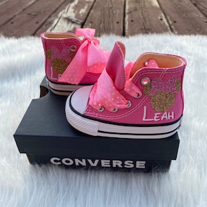 Puede incluir: Zapatillas Converse de ca&ntilde;a alta de color rosa con un dise&ntilde;o de Minnie Mouse de purpurina dorada y una cinta rosa con lunares. Las zapatillas est&aacute;n personalizadas con el nombre "Leah".