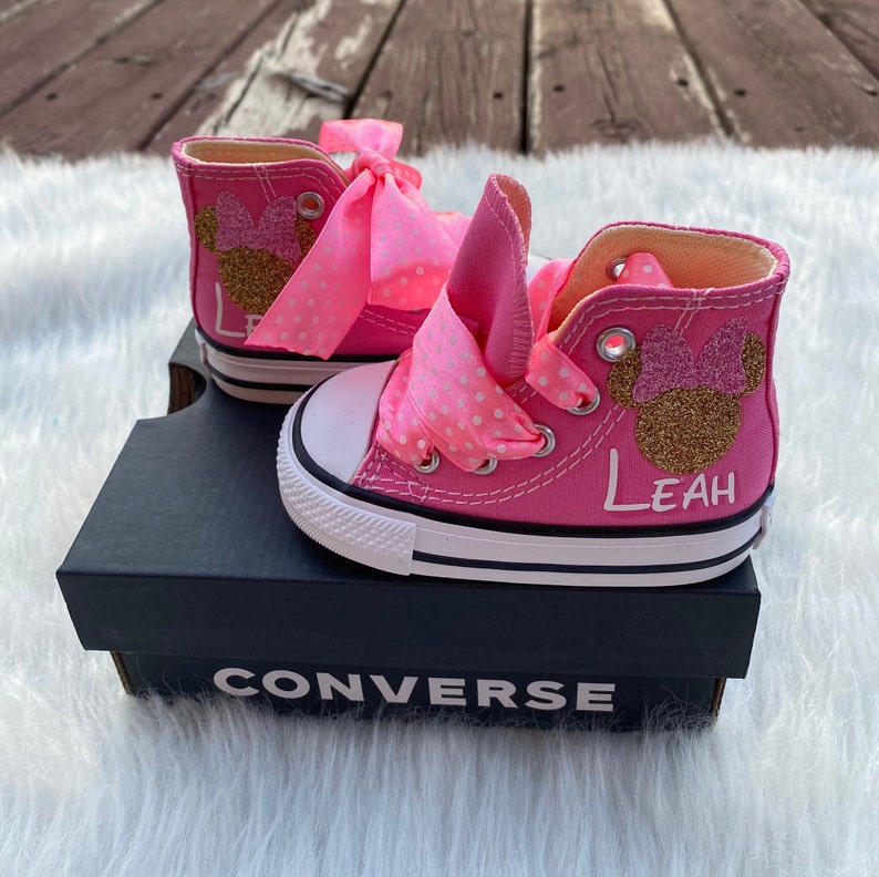 Puede incluir: Zapatillas Converse de ca&ntilde;a alta de color rosa con un dise&ntilde;o de Minnie Mouse de purpurina dorada y una cinta rosa con lunares. Las zapatillas est&aacute;n personalizadas con el nombre "Leah".