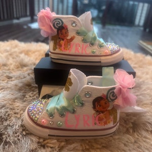 May include: White high-top trainers with a pink bow, green ribbon, and a cartoon character on the side. The trainers are decorated with rhinestones and the name "Lyric" is written on the side.