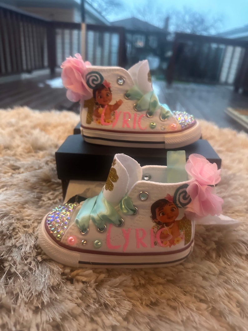 May include: White high-top trainers with a pink bow, green ribbon, and a cartoon character on the side. The trainers are decorated with rhinestones and the name "Lyric" is written on the side.